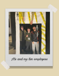 A Polaroid picture taped to a background of three young men in halloween costumes posing for a picture behind a cardboard halloween frame. TEXT: Me and my two employees.